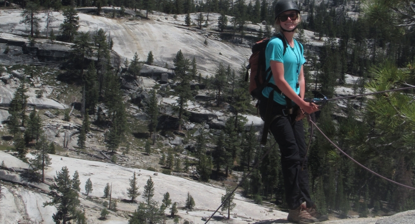 A person wearing safety gear is secured by ropes as they pause climbing a rock face to smile for the photo.